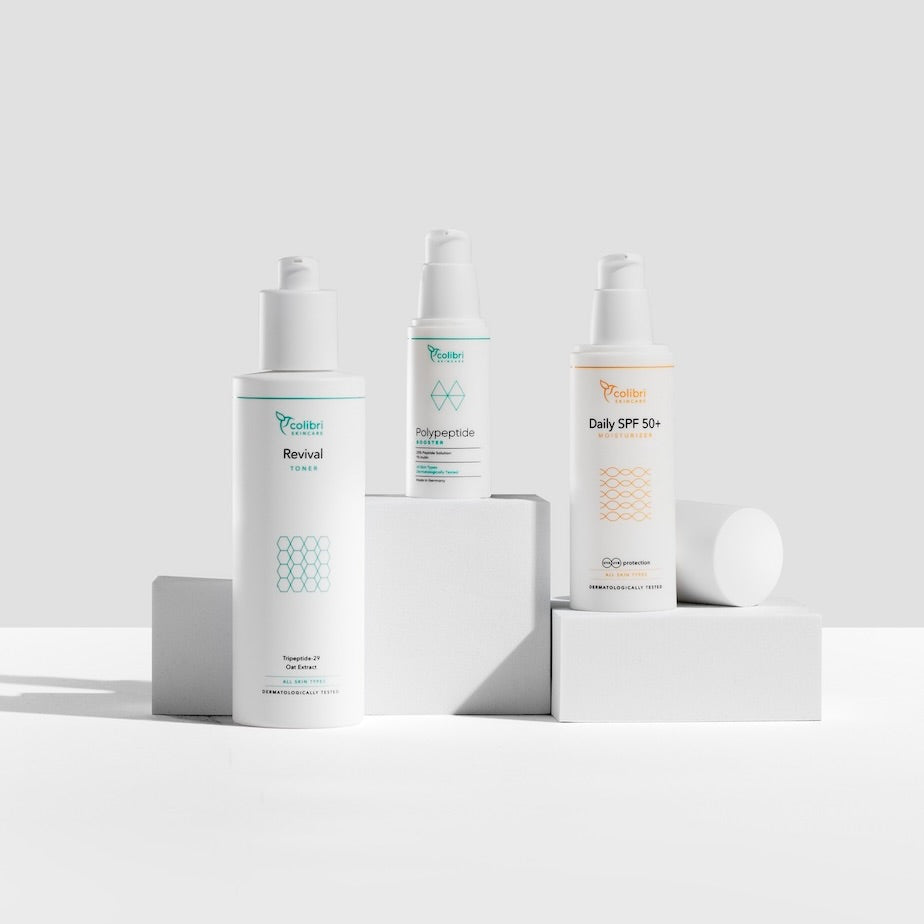 Drei Kolibri Skincare Produkte auf weißen Blöcken vor grauem Hintergrund.