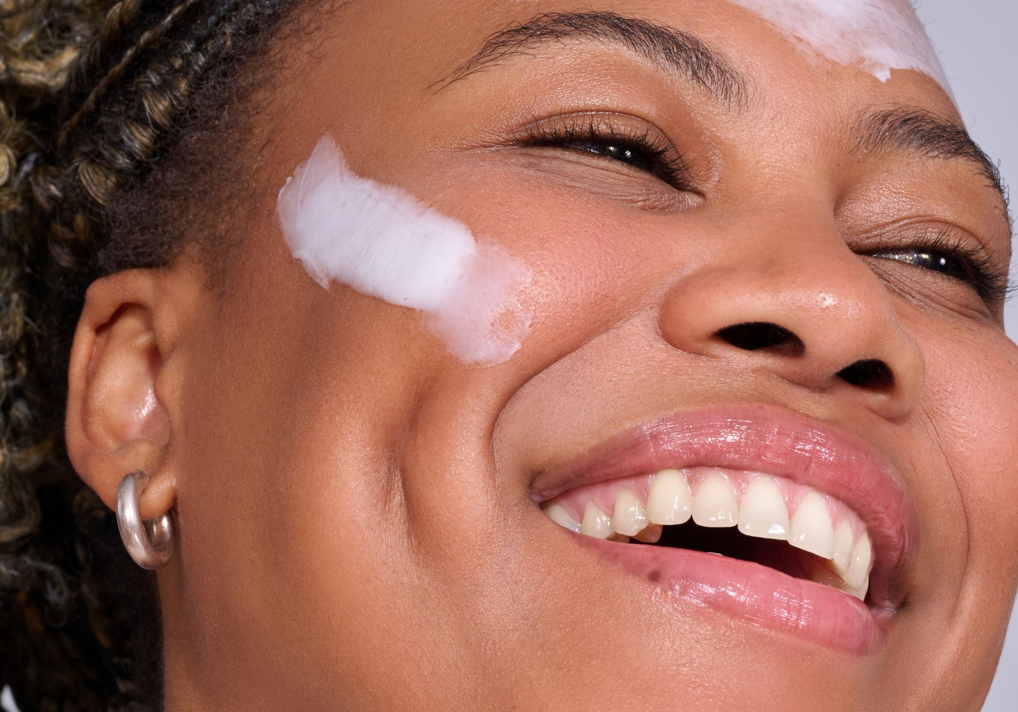 Lachende Person mit Creme auf der Wange, Nahaufnahme des Gesichts mit sichtbaren Zähnen und Ohrring.