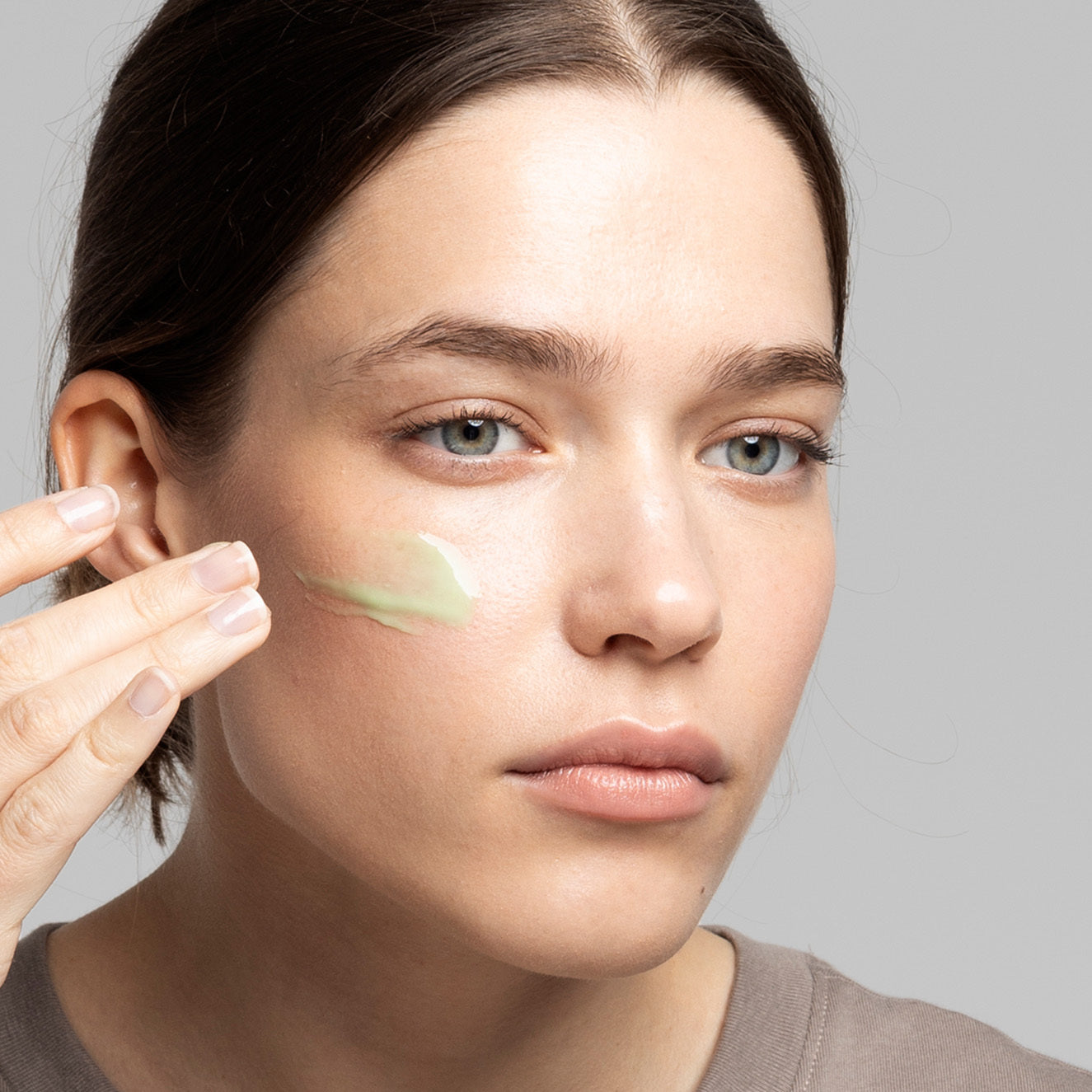 Frau trägt grüne Gesichtscreme auf die Wange auf, Nahaufnahme vor grauem Hintergrund.