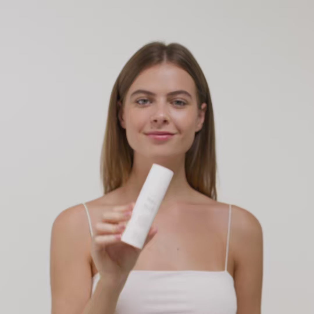 Frau mit langen Haaren hält eine weiße Flasche Kosmetik vor einem neutralen Hintergrund in der Hand.