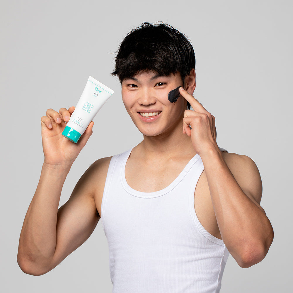 Ein Mann in einem weißen Tanktop trägt eine schwarze Creme auf seine Wange auf und hält eine Tube in der Hand.