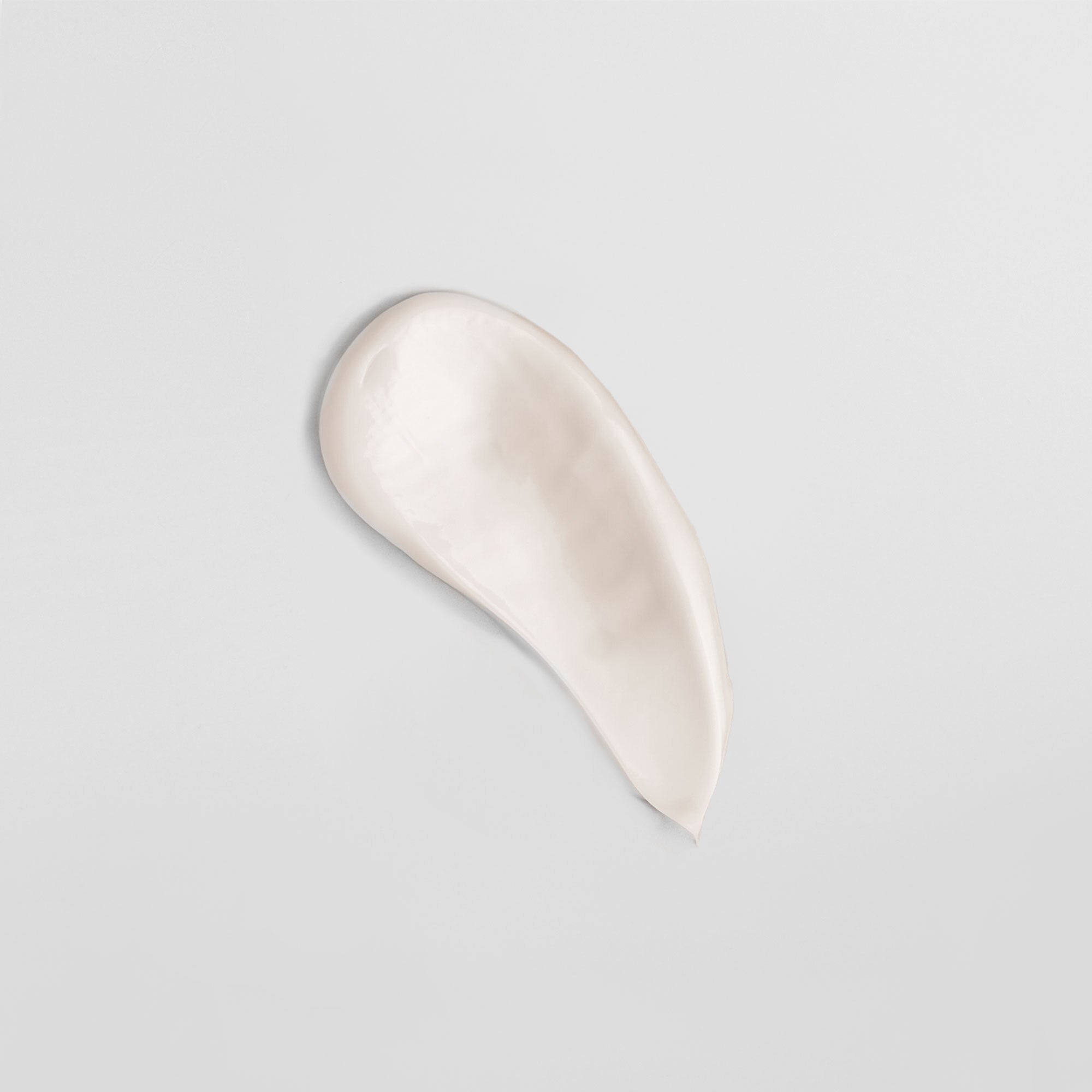 Eine weiße Creme wird in einem langgezogenen Tropfen auf einer neutralen Oberfläche verteilt.